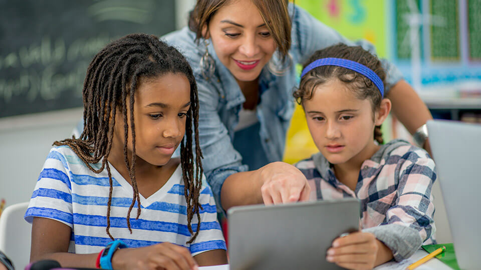 Lerares wijst naar tablet waar twee meisjes mee werken.