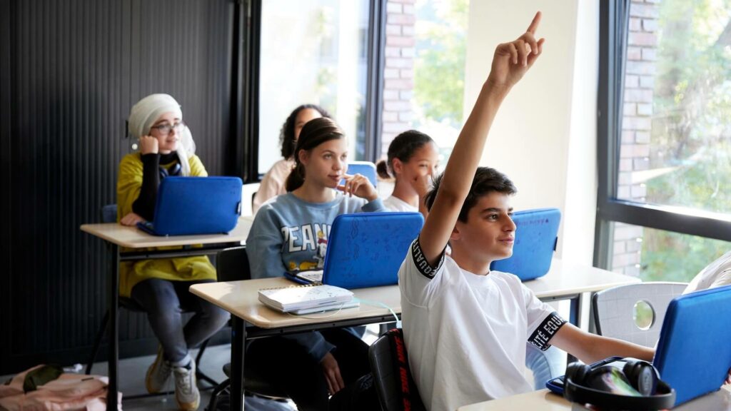 Middelbare schoolleerlingen zitten in een klaslokaal achter hun laptop, een jongen steekt zijn vinger op. 